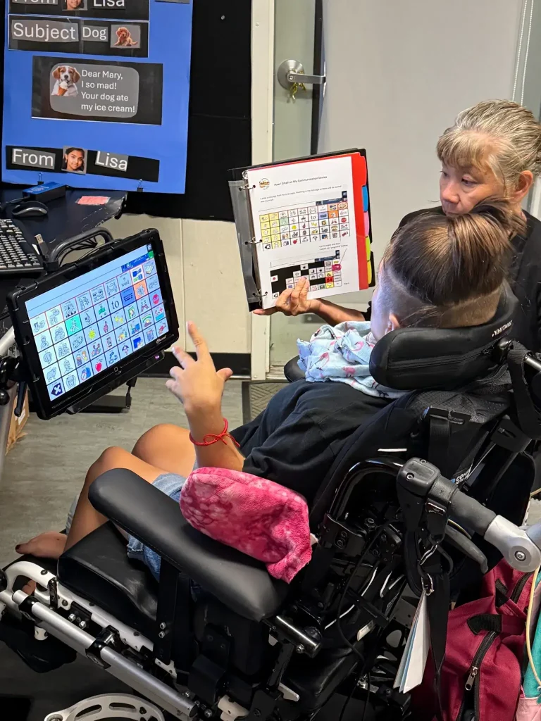 A student in a motorized wheelchair with an AAC device in front of them, is seated next to an adult who is holding up a binder for them to see which contains sample diagrams of AAC device layouts on the shown page