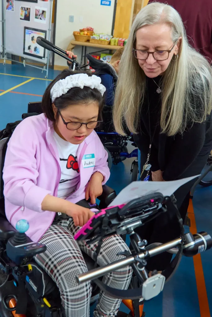 An adult standing and leaning over the shoulder of a child in a motorized wheelchair in order to see what the child is pointing out on their AAC device
